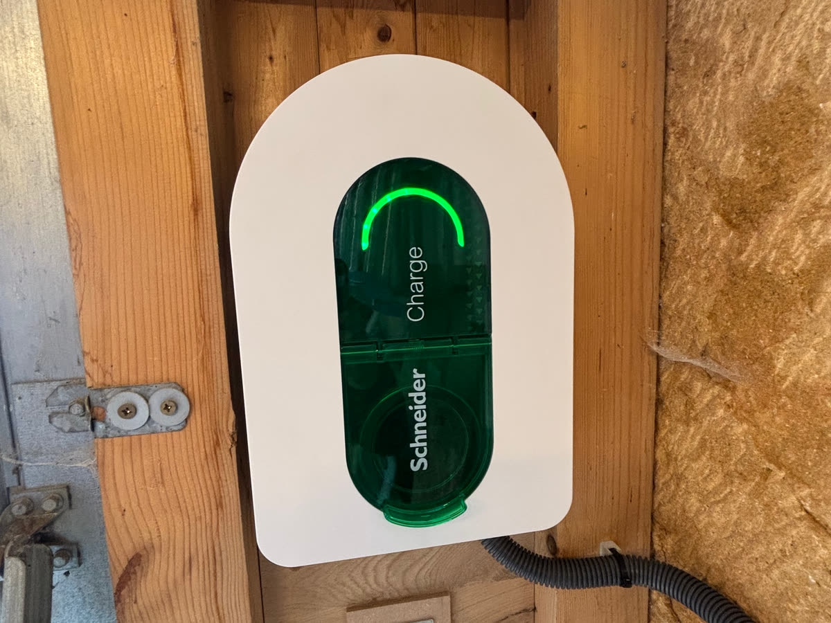 Borne de recharge murale Schneider blanche à façade verte, voyant allumé, fixée sur une ossature en bois, sa gaine d’alimentation partant vers la droite.