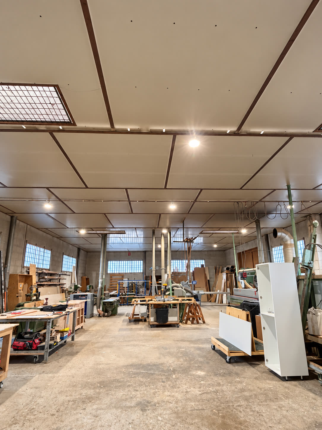 Vue de l’entrepôt de menuiserie en hauteur : grand plafond à panneaux clairs ponctué de luminaires ronds allumés, l’atelier et ses machines en dessous.